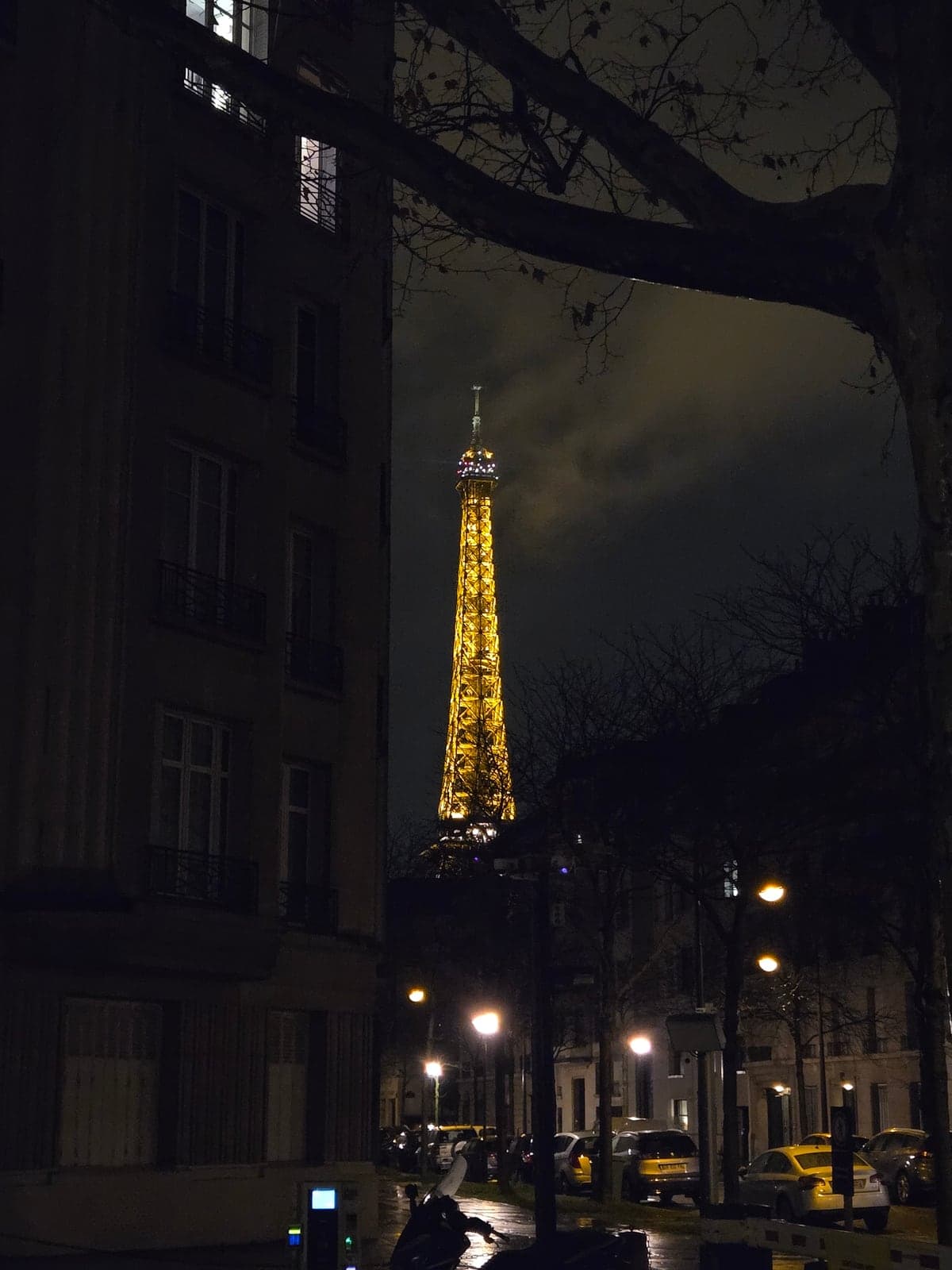 The illuminated Eiffel Tower seen between dark buildings.